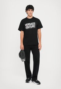 Svart t-skjorte med "VERSACE COUTURE"-logo i hvitt, kombinert med svarte jeans og svarte sneakers, holder en grå caps med samme logo.