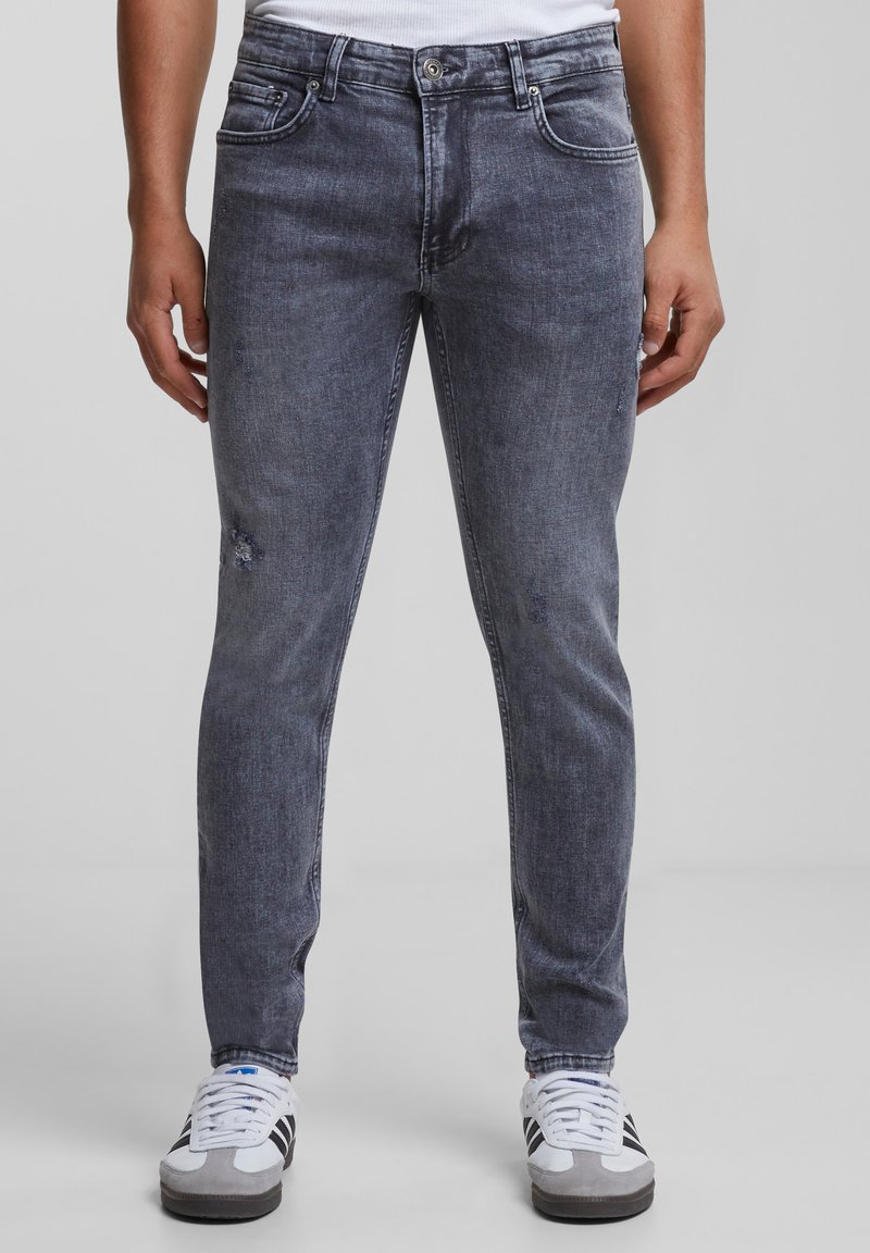 Slim-fit sive traperice s laganim istrošenjima, dizajnom s pet džepova i dugmetom za kopčanje, uparen s bijelim i sivim tenisicama.