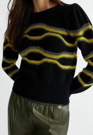 Vrouw die een zwarte trui draagt met gele en grijze golvende strepen en groene hoge taillebroek met een klein metalen logo.