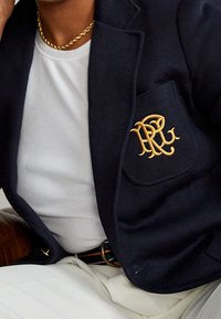 Personne portant un blazer bleu marine avec un logo brodé en or sur la poche, une chemise blanche côtelée, un pantalon blanc et un collier chaîne en or.