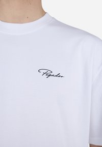 Camiseta blanca de manga corta hecha de algodón con cuello redondo y un pequeño logo negro "Pegador" impreso en el lado izquierdo del pecho.