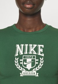 Detailní záběr na osobu v zeleném tričku s bílým logem Nike, nápisem "1972" a textem "Oregon" vytištěným na přední straně.