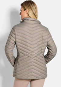 Vrouw met blond haar die een taupekleurige gewatteerde jas draagt, gezien vanaf de achterkant, staand tegen een effen lichte achtergrond.
