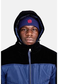 Giacca zip-up navy e nera con colletto alto, realizzata in tessuto liscio. Indossata con un berretto di lana viola con logo di una rosa rossa.