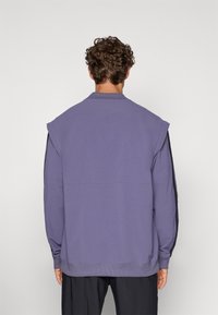 Felpa viola con scollo rotondo, spalle scese e polsini a costine. Il tessuto ha una texture morbida con pannelli laterali a contrasto.