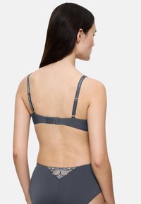 Sieviete ar garām tumšām matiem, ģērbta pelēkā dabiska krūštūrī un augsta jostasvietas apakšveļā ar ziedu mežģīņu detaļām, stāv ar muguru pret balto fonu.