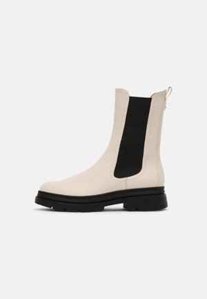 Cremefarbener Leder-Stiefel mit elastischem schwarzem Seitenpanel und klobiger schwarzer Sohle, seitlich auf hellem, einfarbigem Hintergrund gesehen.