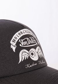 Gorra negra con gráfico blanco que muestra alas y un ojo, el texto dice "Club de Motocicletas Von Dutch Kustom Kulture." La textura parece suave.