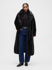 Manteau en fausse fourrure noire avec un large col, porté sur un haut noir et un jean bleu à jambes larges. Texture lisse avec une finition brillante.