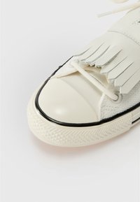 Baskets blanches avec semelle en caoutchouc noir et bout contrastant. Présentent un dessus texturé, des détails à franges et des lacets plats à travers les œillets.