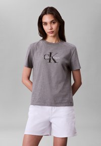 Nő szürke Calvin Klein pólóban és fehér rövidnadrágban, kezét a hát mögött tartva, egyszínű szürke háttér előtt áll.