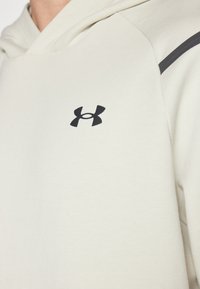 Hellbeige Hoodie aus weichem Stoff, mit einem schwarzen Under Armour-Logo auf der Brust und einem kontrastierenden schwarzen Streifen auf der Schulter.