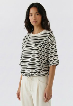 Vrouw met lang donker haar draagt een losse zwart-wit horizontaal gestreepte blouse met een trekkoord aan de onderkant en lichtgekleurde broek tegen een effen achtergrond.