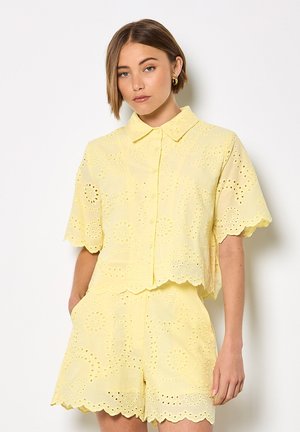 Junge Frau mit kurzen Haaren trägt ein gelbes Kurzarmhemd aus Lochstickerei und passende Shorts, steht vor einer weißen Wand.