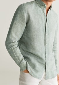 Man in een lichtgroene linnen overhemd met lange mouwen en witte broek, hand in de zak, voor een neutrale achtergrond.