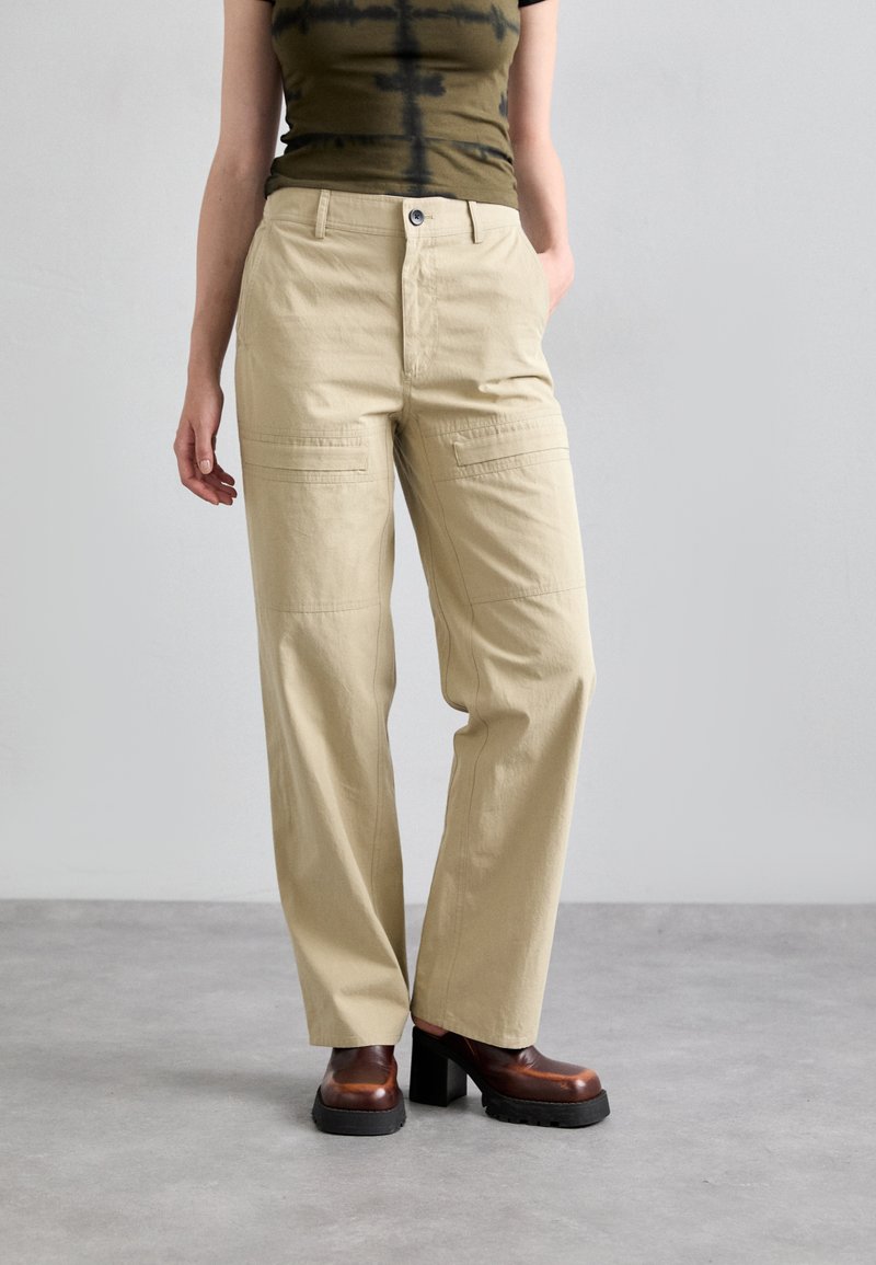 Beige cargo pants with a loose fit, featuring front pockets and a button closure. Worn with a patterned olive tank top and chunky heels.