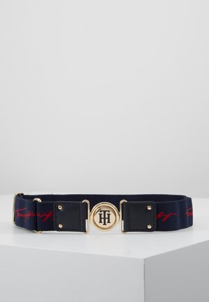 Ceinture en tissu bleu marine avec écriture rouge, boucle ronde dorée avec logo noir "TH", et accents en cuir noir avec rivets dorés.