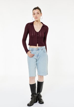 Vrouw draagt een gebreid bordeauxrood crop top met knopen, lichtblauwe denim shorts tot op de knie, zwarte kniehoge laarzen en grote oorringen.