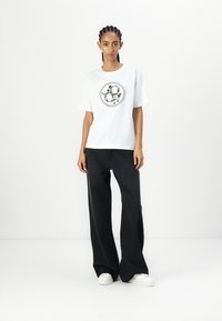 White cotton T-shirt with a circular black graphic design on the front, paired with wide-leg black jeans and white sneakers.