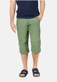 Grüne Capri-Hosen aus strukturiertem Stoff, mit seitlichen Taschen und umgeschlagenen Säumen, kombiniert mit dunklen Sandalen.