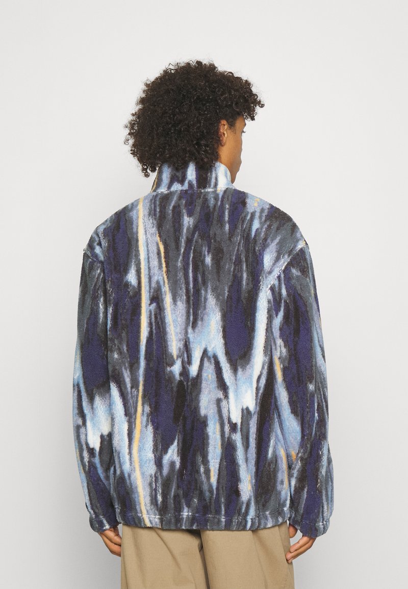 Giacca in pile caratterizzata da un motivo marmorizzato blu, nero e giallo, colletto alto e vestibilità ampia, con cuciture visibili e tessuto testurizzato.