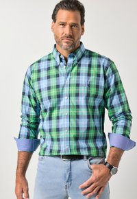 Karierte Hemdjacke in Grün, Blau und Marineblau mit langen Ärmeln und geknöpften Manschetten. Kombiniert mit hellen Denim-Jeans und einer schwarzen Uhr.