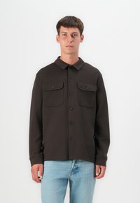 Camisa marrón de botones, hecha de un material texturizado, con cuello, dos bolsillos frontales y botones negros, llevada sobre unos jeans de color azul claro.