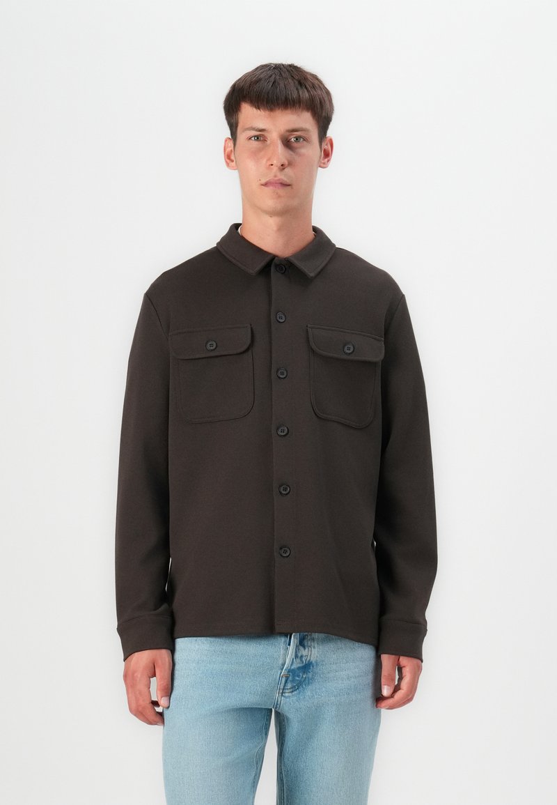 Chemise marron à boutons en matière texturée, dotée d'un col, de deux poches avant et de boutons noirs, portée sur un jean bleu clair.