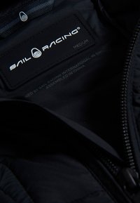 Sort jakke indvendig med et logo mærke "SAIL RACING" i hvid tekst; inkluderer størrelse "MELLEM". Lynlås og blødt quiltet stof synligt.