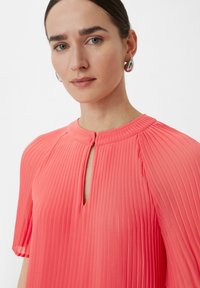 Zgubiona koralowa sukienka z dekoltem w kształcie łezki i krótkimi rękawami. Wykonana z lekkiego materiału, charakteryzującego się gładką fakturą i subtelnym połyskiem.