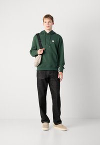 Felpa verde con cappuccio, tasca frontale e toppa con logo, abbinata a jeans scuri e sneakers beige. Il modello tiene una borsa grigia chiara.