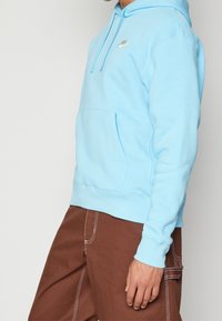 Persona vestida con una sudadera Nike azul claro con bolsillo frontal y pantalones marrones con costuras blancas, de pie contra un fondo blanco liso.