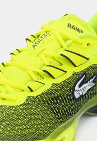 Fotbalové boty Lacoste AGLT23 Ultra mají jasně žlutý síťovaný svršek s černými akcenty, texturovaným designem a výrazným bílým logem.
