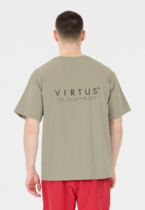 Mand med kort mørkt hår iført en løs lyse olivengrøn T-shirt med teksten "VIRTUS" og røde shorts, set bagfra.