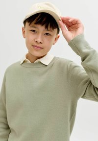 Jeune personne portant une casquette beige, un pull vert clair et une chemise écrue à col, ajustant la casquette de la main droite.