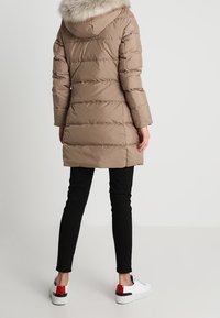 Béžová péřová bunda s kapucí lemovanou kožešinou, prošívanou strukturou a dlouhými rukávy, v kombinaci s černými kalhotami a bílými teniskami s červenými akcenty.