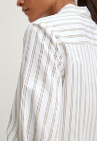 Witte blouse met verticale beige strepen, met een zachte textuur en een relaxed fit. Detail toont de schoudernaad en kraag.