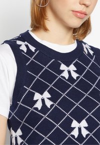 Femme portant un gilet en tricot bleu marine avec un nœud blanc et un motif en losange sur une chemise blanche à manches courtes, montrant une partie de son visage et de son épaule.