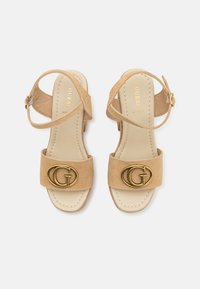 Sandales à talon en suède beige avec un bout ouvert, une bride de cheville et un logo "G" doré bien visible sur le devant. Semelle intérieure lisse et couture minimale.