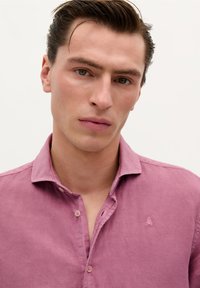 Cămașă din in roz deschis cu guler button-down, având un mic logo brodat pe piept; textură moale cu o ușoară strălucire.