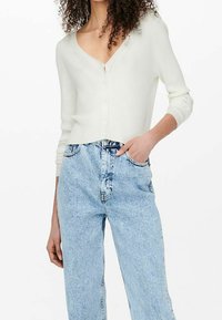 Kobieta w białym prążkowanym kardiganie zapinanym na guziki z dekoltem w serek oraz w jasnoniebieskich jeansach z wysokim stanem i efektem "acid-wash", z jedną ręką w kieszeni.