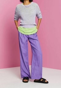 Osoba ubrana w lawendowy sweter dzianinowy na jasnoszarym koszuli, luźne fioletowe spodnie i czarne sandały na ulicy, stojąca na tle różowej ściany.