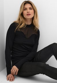 Črna dolga majica z dolgimi rokavi in prosojnim mrežastim predelom, ki ima valovit vzorec. Ujemajoče se z teksturiranimi črnimi leginami z vzorcem v krovnični tematik.