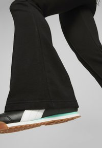 Svarta utsvängda byxor med en slät textur, kombinerat med svarta sneakers med vita ränder och en mintgrön sula.