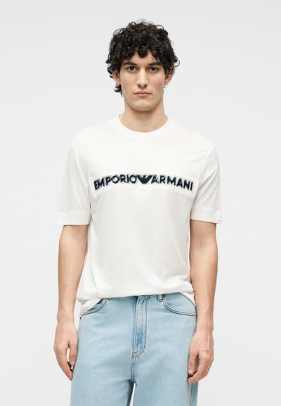 Jeune homme aux cheveux bouclés portant un t-shirt blanc Emporio Armani et un jean bleu clair, debout devant un fond uni.