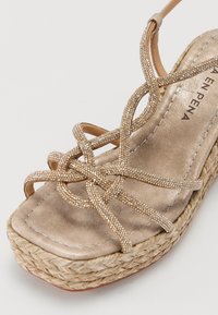 Sandale en wedge dorée avec des brides croisées ornées de petits strass. Semelle texturée ressemblant à de la paille tissée. Semelle intérieure lisse et métallique.