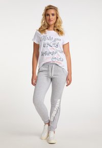 Weißes Baumwoll-T-Shirt mit bedrucktem Text in verschiedenen Schriftarten. Hellgraue Sweatpants mit seitlichem Textdetail. Weiße Sneaker runden das Outfit ab.