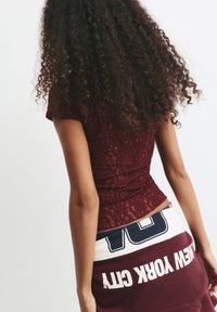 Frau mit lockigem Haar, die ein bordeauxfarbenes Spitzenoberteil und marineblaue Jeans trägt, während sie einen bordeauxfarbenen Sweatshirt mit dem Text "NEW YORK CITY" hinter sich hält.