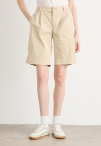 Pantaloni corti beige in cotone con vestibilità rilassata, dotati di pieghe, risvolti e tasche laterali. Abbinati a sneakers bianche e calzini.