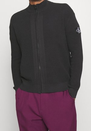 Hombre con suéter negro de cremallera completa con parche de logo Calvin Klein en la manga y pantalones morados, manos en los bolsillos, sobre fondo liso.